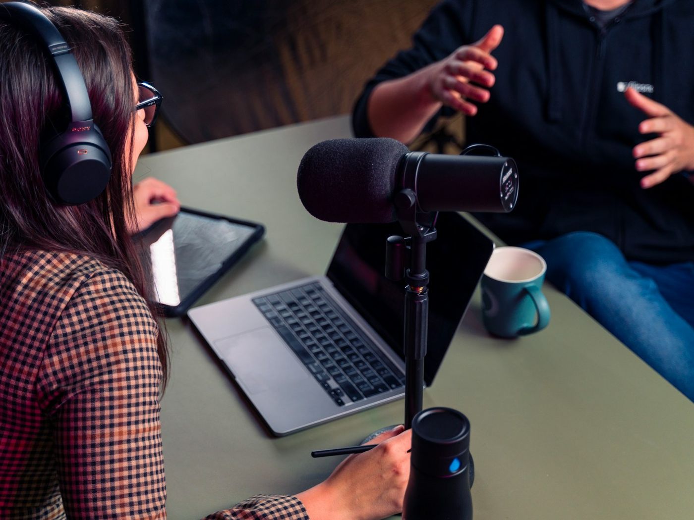 Two people recording a podcast at a table, one wearing headphones and using a laptop
